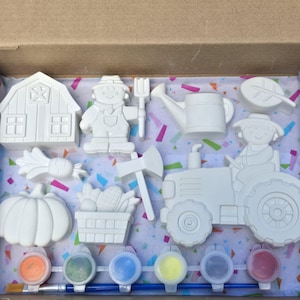 May include: A set of white ceramic figures including a barn, a scarecrow, a watering can, a leaf, a pumpkin, a basket of corn, an axe, and a tractor. The set includes six small paint pots in various colors and a blue paintbrush for painting.