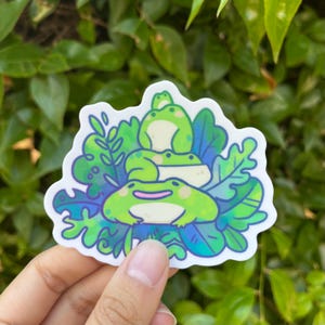 May include: A sticker featuring three cartoon frogs stacked on top of each other, surrounded by green and blue leaves. The frogs are light green with pink accents. The sticker has a white border.