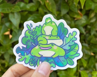 May include: A sticker featuring three cartoon frogs stacked on top of each other, surrounded by green and blue leaves. The frogs are light green with pink accents. The sticker has a white border.