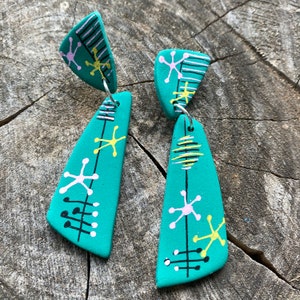 Puede incluir: Un par de pendientes colgantes de color azul turquesa con formas geométricas y detalles en blanco, negro y amarillo. Los pendientes están hechos de arcilla polimérica y tienen un diseño moderno de mediados de siglo.