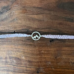 May include: A purple braided bracelet with a silver charm featuring a mountain silhouette.