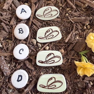 May include: Four white circular markers labeled A, B, C, and D, paired with four off-white, smooth, flat pieces, each featuring a dark brown line drawing of a cowboy hat. The items are arranged in a vertical column on a bed of brown wood mulch.