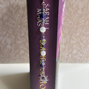 May include: A metal bookmark featuring a series of purple, iridescent, and clear crackle glass beads strung on a silver wire, ending with a silver sun-shaped charm with a small purple center, attached to the spine of a purple book.