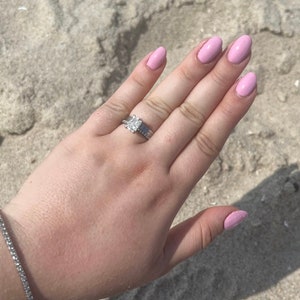 May include: A hand with light pink nail polish and a diamond engagement ring. The ring has a square-shaped diamond with a silver band.