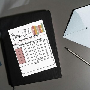 May include: A white Book Club monthly reading tracker with a colorful stack of books graphic. The tracker includes spaces to record the number of pages read each day of the week, with sections for the month, week of, total pages, and notes. It rests on a black notebook.