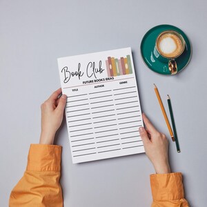 May include: A white Book Club planner with lines for title, author, and genre, held by hands. The planner has the words "Book Club" and "Future Books Ideas" printed on it. A teal cup of coffee and two pencils are on the gray surface.