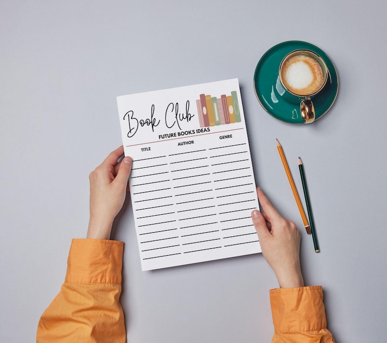 May include: A white Book Club planner with lines for title, author, and genre, held by hands. The planner has the words "Book Club" and "Future Books Ideas" printed on it. A teal cup of coffee and two pencils are on the gray surface.