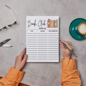 May include: A white Book Club Book Log notepad with the title and book log details printed in black. A person is holding a pencil and writing on the notepad. A pair of gold scissors, glasses, and a cup of coffee are also on the table.