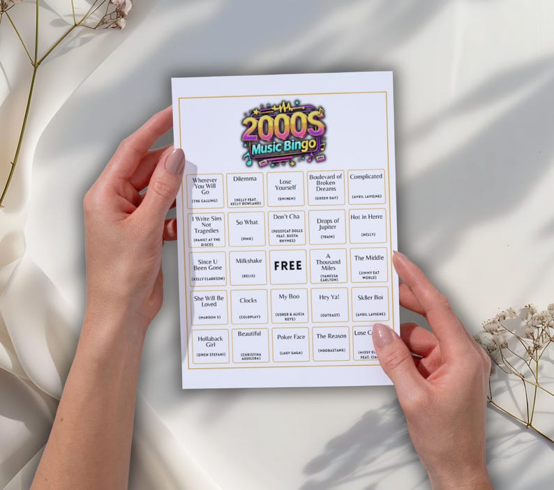 May include: A white bingo card with a gold border is held by two hands. The card is titled "2000s Music Bingo" and features a grid of song titles. The word "FREE" is in the center of the card.