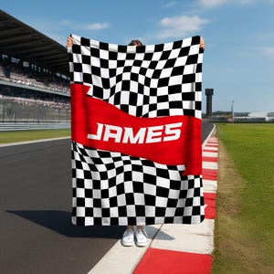 May include: A black and white checkered blanket with a red banner across the center. The word "JAMES" is printed in white on the red banner. The blanket is held up in front of a race track.