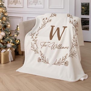 May include: A cream-colored throw blanket featuring a large brown letter W centered within a delicate floral wreath design. Below the letter, the text The Williams is printed in a brown script font, creating a personalized decorative accent.