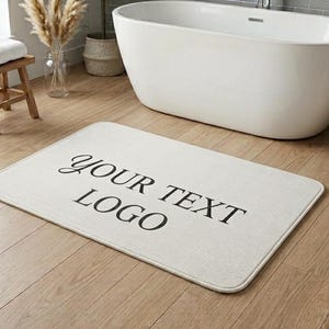 May include: A rectangular off-white bath mat featuring the black text YOUR TEXT LOGO centered on the surface. The mat has rounded corners and a soft, textured appearance, positioned flat on a light brown wooden floor in a bathroom setting.