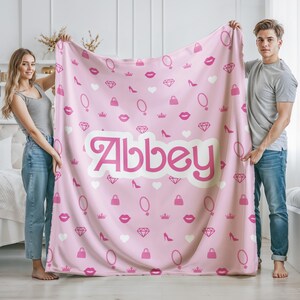 May include: Pink fleece blanket with a white background and a repeating pattern of pink lips, crowns, diamonds, purses, and necklaces. The name "Abbey" is written in a large, pink, cursive font.