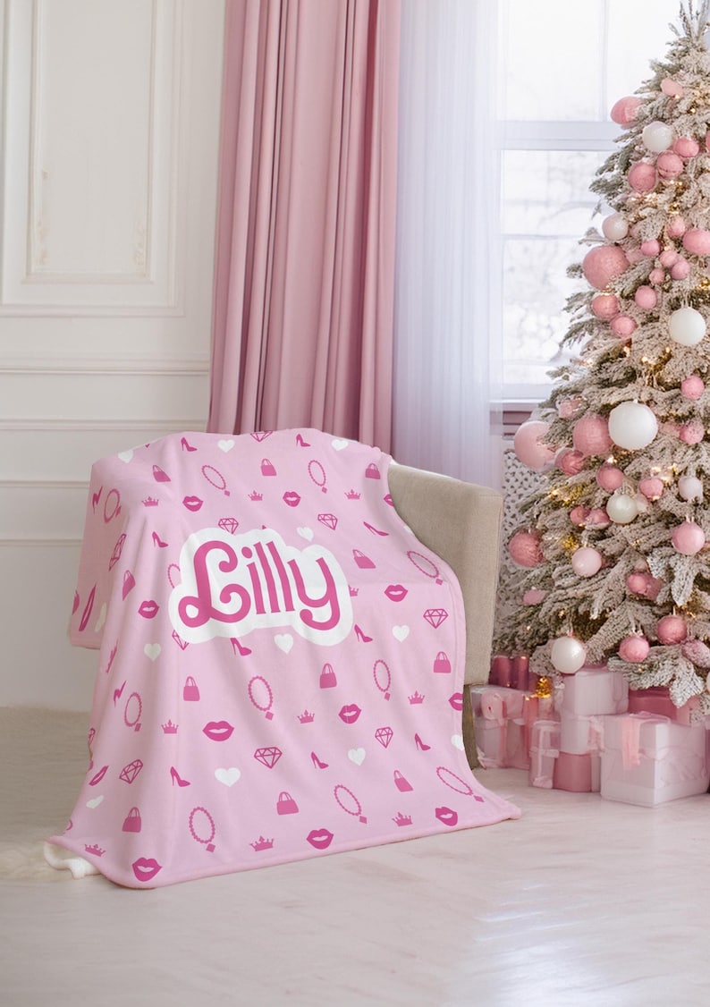 May include: Pink fleece throw blanket with a white heart and the name "Lilly" in a pink and white font. The blanket has a repeating pattern of pink lips, purses, crowns, high heels, and necklaces.