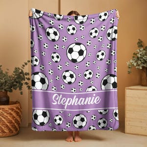 May include: A purple blanket featuring a pattern of soccer balls in various sizes. The blanket has a lavender band with the name "Stephanie" in white script. The soccer balls are black and white.