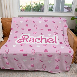 May include: Pink fleece throw blanket with a white  "Rachel" name in a cursive font. The blanket has a repeating pattern of pink lips, diamonds, crowns, purses, high heels, and mirrors.