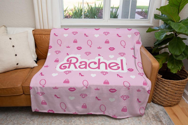 May include: Pink fleece throw blanket with a white  "Rachel" name in a cursive font. The blanket has a repeating pattern of pink lips, diamonds, crowns, purses, high heels, and mirrors.