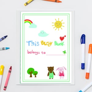 May include: A white framed page with the text "This Busy Book belongs to" and illustrations of a rainbow, sun, cloud, heart, trees, bear, and bunny. Crayons and markers surround the page.