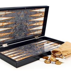 May include: A black backgammon set with a marble-like design in shades of gray, white, and gold. The set is open, revealing the playing surface. A tan drawstring bag spills out game pieces and dice.