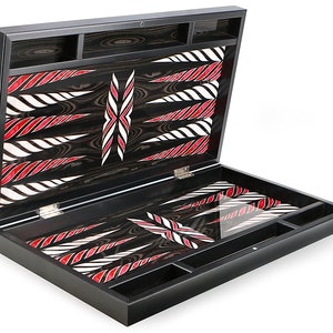 Puede incluir: Un juego de backgammon de madera negra con un diseño de incrustaciones rojas y blancas. El juego está abierto y la superficie de juego es visible. El juego tiene compartimentos para guardar las piezas del juego.
