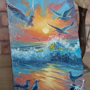 May include: A painting of a seascape with a bright orange and yellow sunset, blue waves crashing on the shore, and seagulls flying overhead. The painting is on a small canvas with a wooden frame.