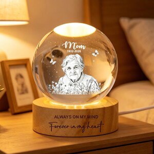 Può includere: Una lampada a sfera di cristallo luminosa con un ritratto di donna, la parola "Mom" e gli anni 1932-2026. La lampada è su una base di legno con l'iscrizione "Always on my mind, Forever in my heart."