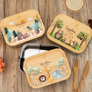 Puede incluir: Tres fiambreras personalizadas de bambú con interiores de acero inoxidable. Cada tapa presenta una escena diferente de animales del bosque con nombres y fechas. Incluye un tenedor y un cuchillo de madera.