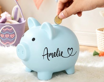 Peut inclure: Une tirelire bleu clair avec le prénom Amelie écrit en lettres cursives noires accompagné d'un petit cœur. Le cochon en céramique possède deux yeux noirs circulaires et une fente sur le dessus pour insérer des pièces de monnaie.