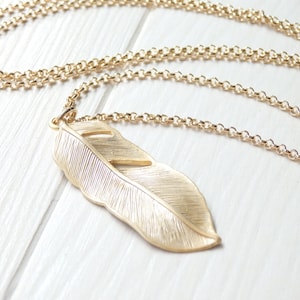 May include: A gold-coloured necklace with a large feather pendant. The feather pendant has detailed veining and a cutout design. The chain is a delicate, linked style. The necklace is displayed on a white surface.