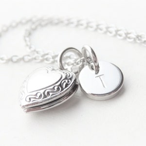May include: A silver necklace with a heart-shaped locket and a circular charm engraved with the letter "T". The locket has decorative detailing. The necklace is shown on a white background, highlighting the chain.