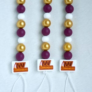 Op de afbeelding: Drie kralen lanyards met een herhalend patroon van gouden, paarse en witte kralen. Elke lanyard heeft een witte vierkante bedel met het logo en de tekst van de Washington Commanders. Witte koorden hangen aan de bedels.
