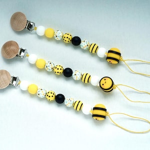 Op de afbeelding: Drie houten klemmetjes voor speenkettingen met gele, witte en zwarte kralen. Elk klemmetje heeft een bij-vormige hanger met een glimlachend gezicht.