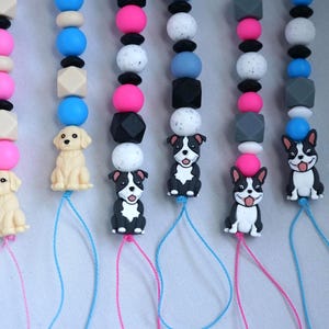 May include: A collection of colorful beaded pacifier clips. Each clip features a different design, including a golden retriever and a black and white dog. The beads are in various colors, including pink, blue, black, and white.