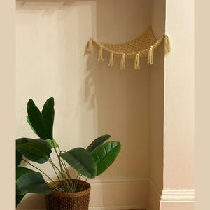 Puede incluir: Una hamaca para gatos beige tejida con borlas cuelga en una esquina. Una planta en maceta con hojas grandes y verdes se encuentra en el suelo debajo. La planta está en una cesta tejida marrón.