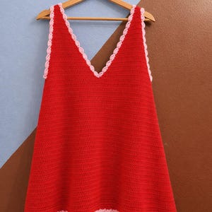 May include: A red crochet top with a V-neck and a scalloped pink trim. The sleeveless top is hanging on a wooden hanger against a two-tone blue and brown wall. The handmade garment is a summer top.
