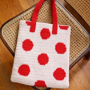 Op de afbeelding: Een handgemaakte gehaakte tote bag met een witte basis en grote rode stippen. De tas heeft rode handvatten en staat op een houten stoel met een geweven zitting. Een stijlvol accessoire.