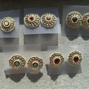 May include: A set of six pairs of round, gold-tone earrings with white pearl accents. Each pair features a different coloured gemstone in the centre: pink, red, green, turquoise, and red.