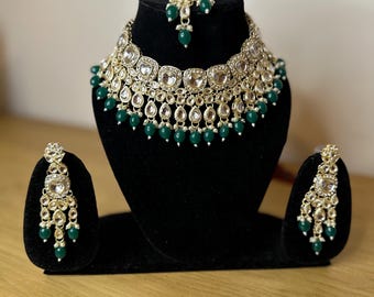 May include: A gold and green Indian bridal jewellery set featuring a necklace, earrings, and a maang tikka. The necklace has a large, round pendant with smaller stones and beads surrounding it. The earrings are long and dangle, with green beads and stones. The maang tikka is a small, ornate piece that sits on the forehead.