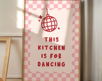 May include: A framed print with a light pink background and a pink and white checkered border. The print features a red disco ball illustration and the text "THIS KITCHEN IS FOR DANCING" in red. The frame is made of light-colored wood.