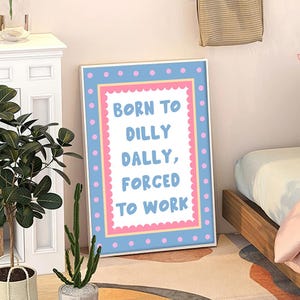 May include: A framed print with the text "BORN TO DILLY DALLY, FORCED TO WORK." The frame has a blue border with pink polka dots and a pink inner border. The background is white with the text in blue.