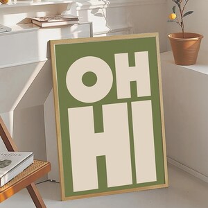 May include: A framed art print with the words "OH HI" in large, bold, off-white letters against a green background. The frame is a light gold color. The artwork is leaning against a white wall.