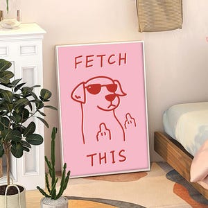 Op de afbeelding: Een ingelijste roze kunstprint met een cartoonhond met een zonnebril en de middelvinger. De rode tekst luidt "FETCH THIS". Het kunstwerk wordt weergegeven in een witte lijst.