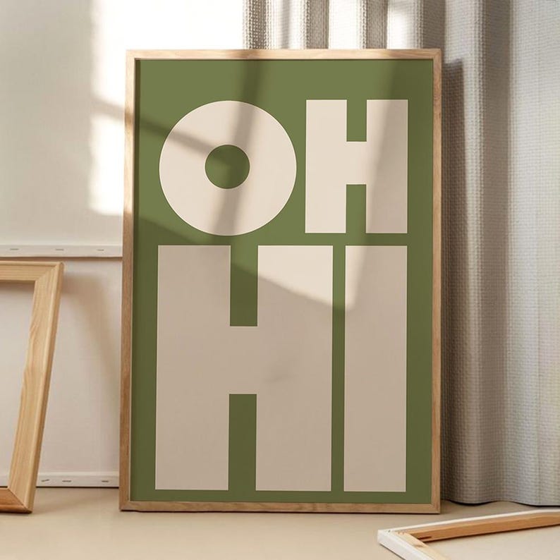 May include: A framed art print with the words "OH HI" in large, bold letters. The background is a muted green, with the letters in a cream color. The frame is a light wood color.