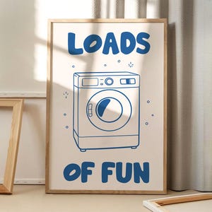 Op de afbeelding: Een ingelijste print met een lichtbeige achtergrond en blauwe tekst met de tekst "LOADS OF FUN". Een blauwe lijntekening van een wasmachine is gecentreerd onder de tekst. De print zit in een lichtbruine houten lijst.