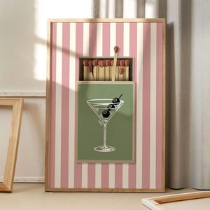 May include: A framed art print featuring a martini glass with olives and a matchbox design. The background has pink and white vertical stripes. The martini glass is on a green background, and the matchbox is open with matches.