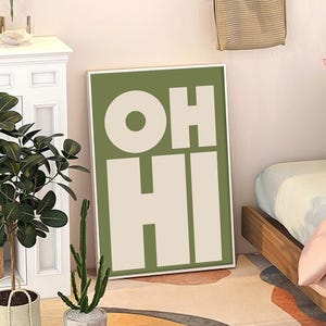 May include: A framed art print with the words "OH HI" in large, bold, white letters against a green background. The artwork is leaning against a wall, with a plant and a bed visible in the background.