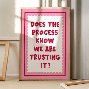 May include: A framed print with the text "DOES THE PROCESS KNOW WE ARE TRUSTING IT?" in red on a white background. The print has a pink scalloped border and is set in a wooden frame. The artwork is displayed indoors.