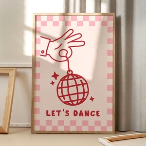 Op de afbeelding: Een ingelijste kunstdruk met een lichtroze achtergrond en een rode illustratie. De illustratie toont een hand die een discobal vasthoudt met de tekst "LET'S DANCE" eronder. De rand heeft een roze en wit geblokt patroon.