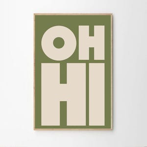 May include: A framed art print with the words "OH HI" in large, bold, cream-colored letters against a solid olive green background. The letters are in a sans-serif font. The frame is a light brown color.