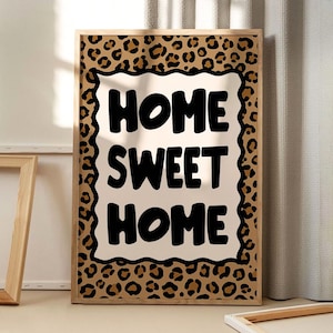 Può includere: Stampa artistica da parete incorniciata con la scritta "HOME SWEET HOME" in grassetto nero. L'opera presenta uno sfondo color crema, un bordo con stampa leopardata e una cornice in legno marrone chiaro.
